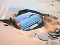 洪水で陥没して空いた道路の穴に車がゆっくりとのみこまれていく様子。